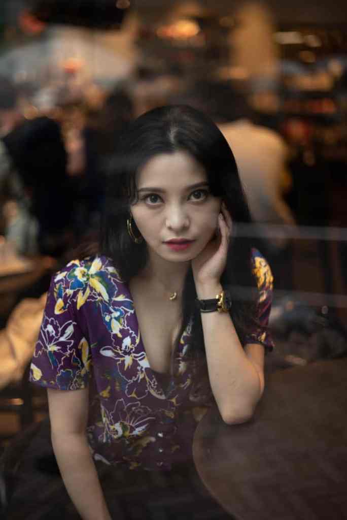 Mulher olhando para a câmera com a mão apoiada no rosto e sentada, ela usa camisa roxa estampada de flores e usa várias joias de ouro e relógio