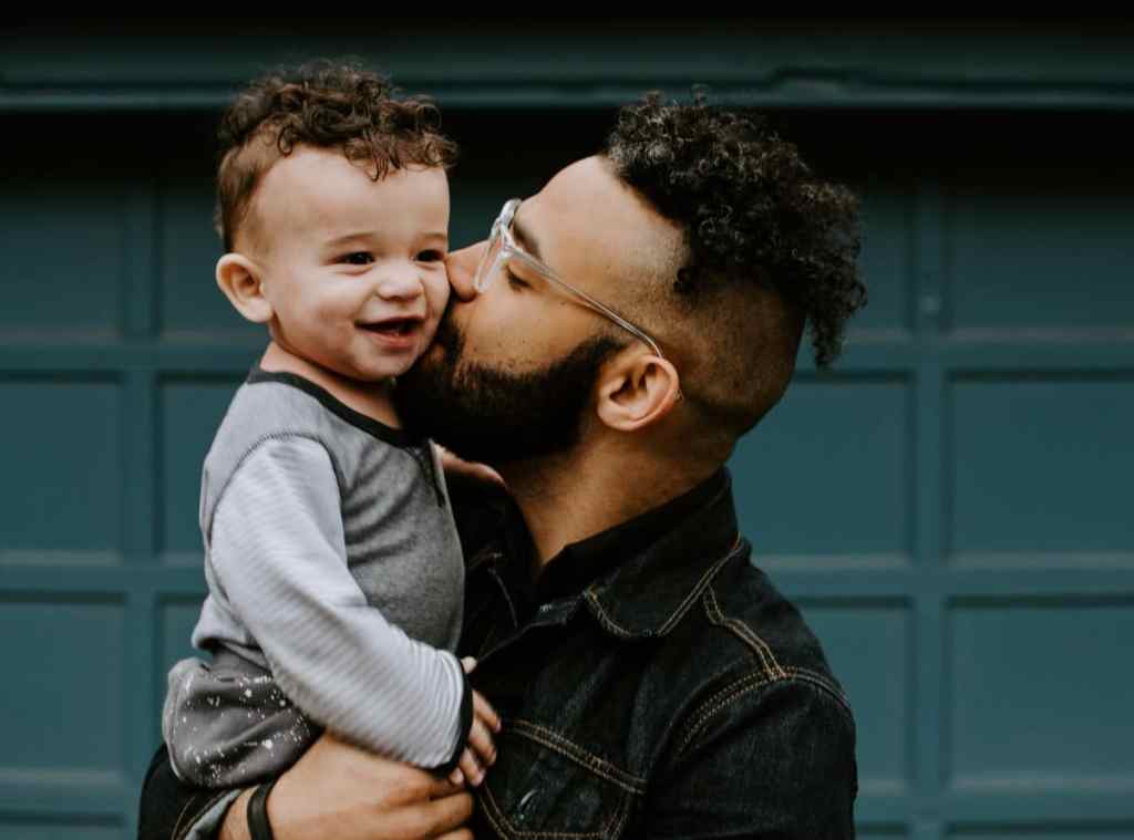 Homem dando beijo na bochecha do filho enquanto o segura no colo. O homem usa óculos, barba e camisa xadrez
