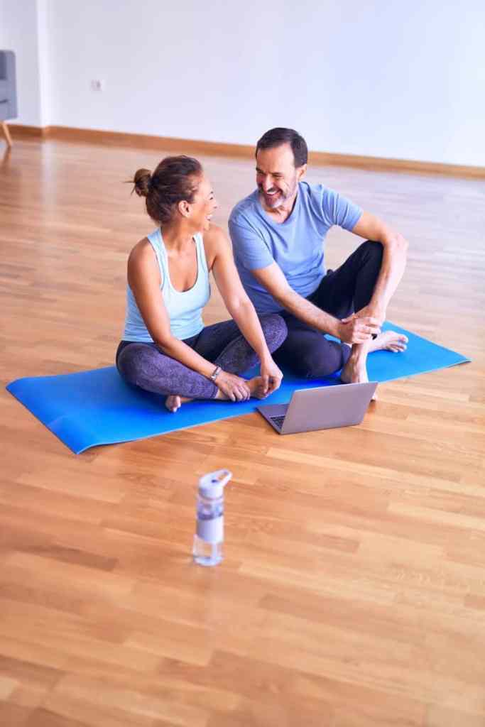 Casal sentado em tapete de yoga sorrindo juntos, em frente eles tem um notebook e uma garrafa de água no chão