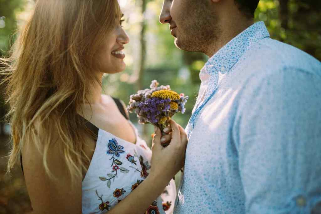 Homem e mulher de frente par ao outro, ela está segurando um pequeno buquê de flores e sorrindo para ele