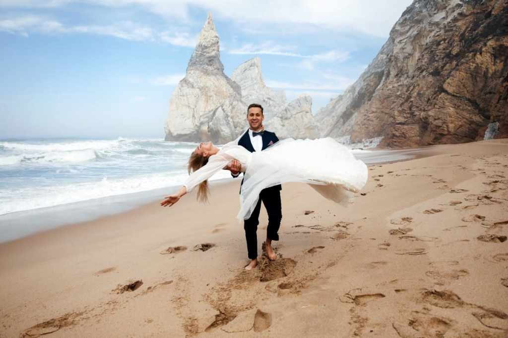 Noivo carregando noiva no colo na praia, ele está descalço