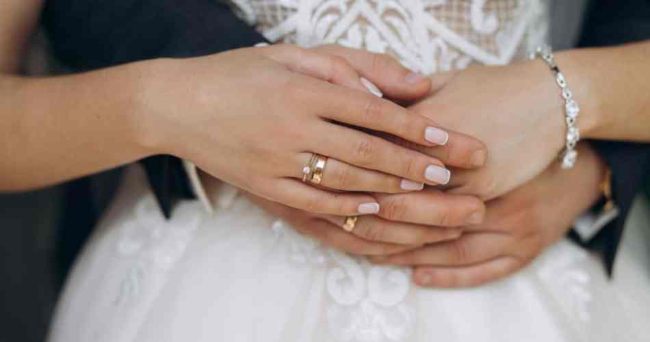 Casal de noivos abraçados, ele está com as mãos rodeadas na cintura dela, e as mãos dela estão por cima onde é possível ver a aliança e o anel de noivado