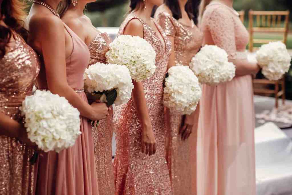 Várias madrinhas de casamento usando vestido rosa bebê com formatos e tecidos diferentes, elas estão em linha no casamento esperando a entrada e segurando o buquê