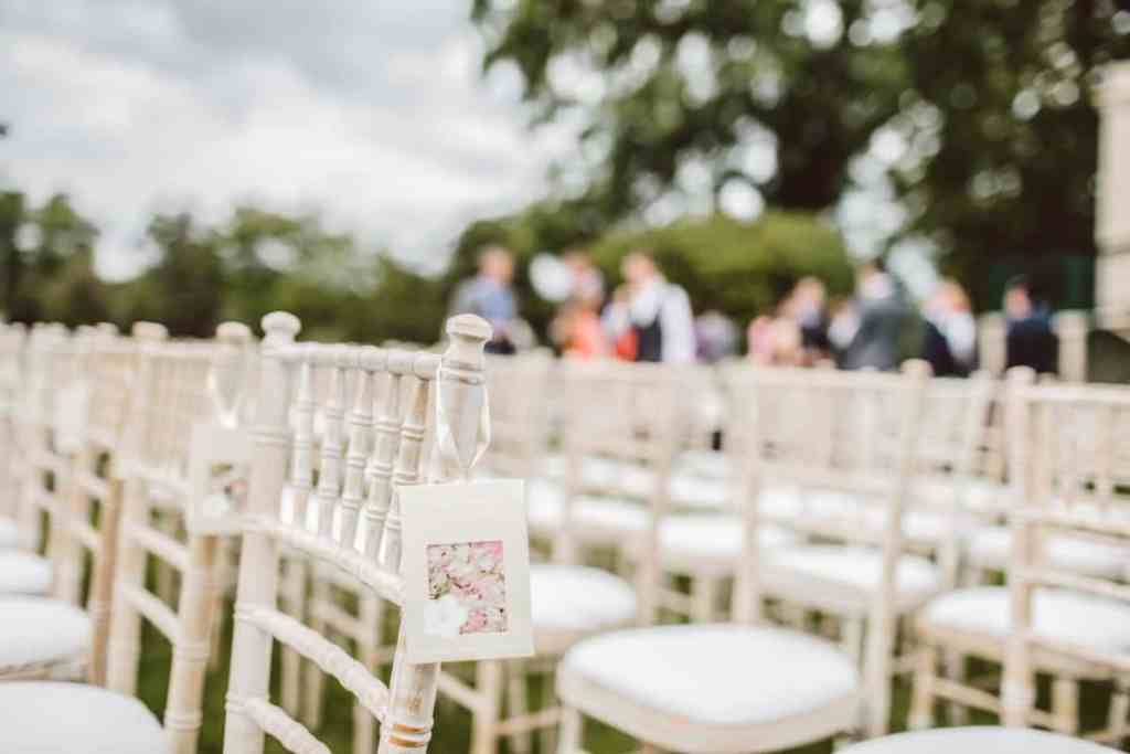 Casamento ao ar livre, a foto foca nas cadeiras vazias do casamento, no fundo desfocado são os convidados chegando
