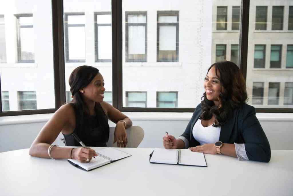 Duas mulheres em um escritório branco fazendo anotações na agenda e olhando uma pra outra sorrindo