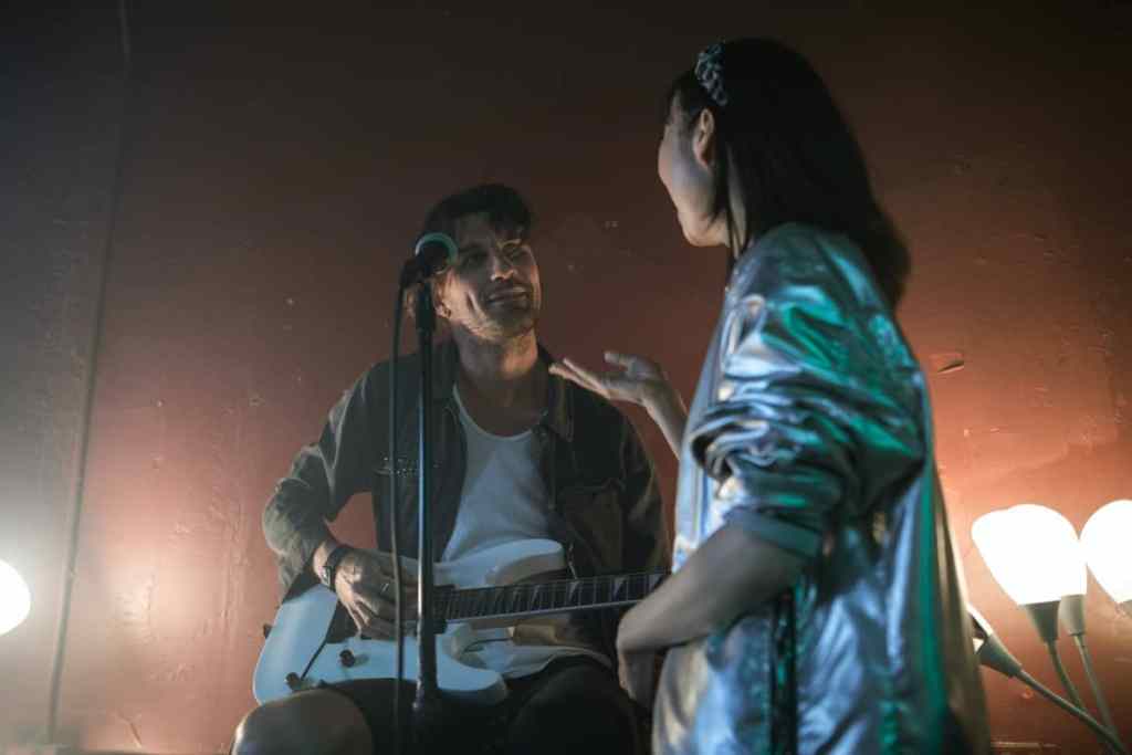 Homem e mulher em palco, ele está sentado segurando uma guitarra e ela em pé em frente ao microfone, eles estão olhando um para o outro