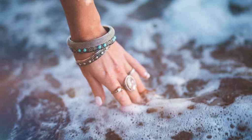 Mulher passando a mão na água do mar, ela está usando pulseiras e anéis