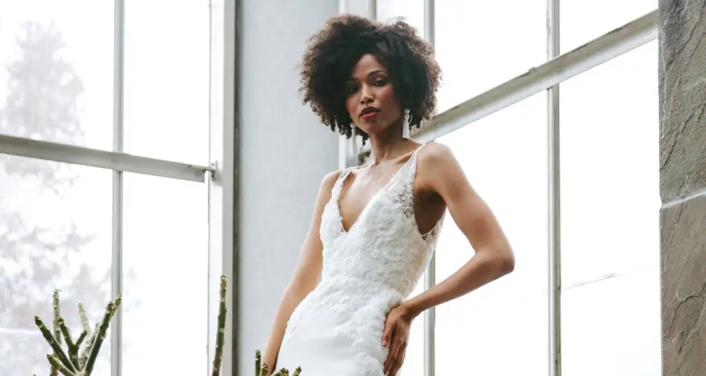 Noiva negra com cabelo crespo curto solto, ela usa vestido de noiva de alça branco e está posando com mão na cintura