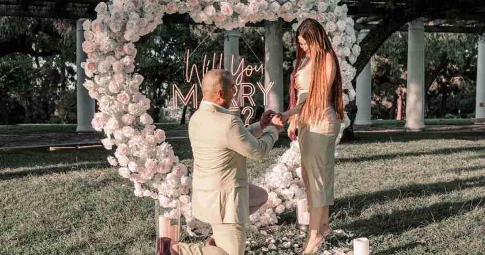 Homem fazendo pedido de casamento em frente a arco de fores em formato de coração
