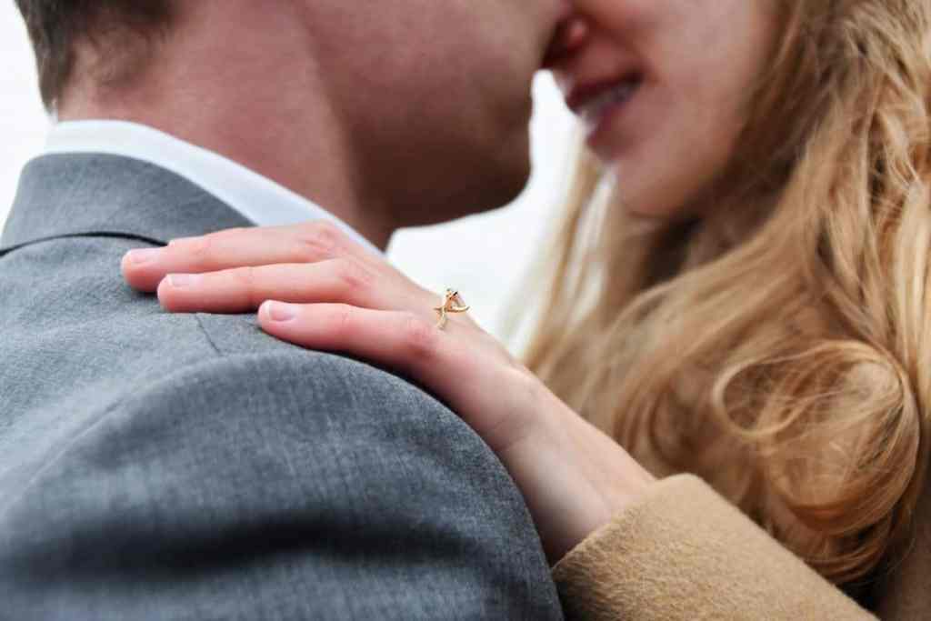 Homem e mulher com rosto bem próximos, a foto está focando na mão dela que tem um anel de noivado em ouro