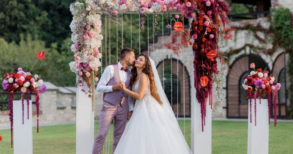 Noivo indo beijar bochecha da noiva, é um casamento ar livre, atrás deles tem um altar com flors na cor marsala