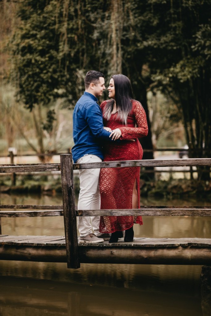 Casal em cima de ponte de madeira em parque, ele está abraçando ela pela cintura, ela usa vestido rendado vermelho