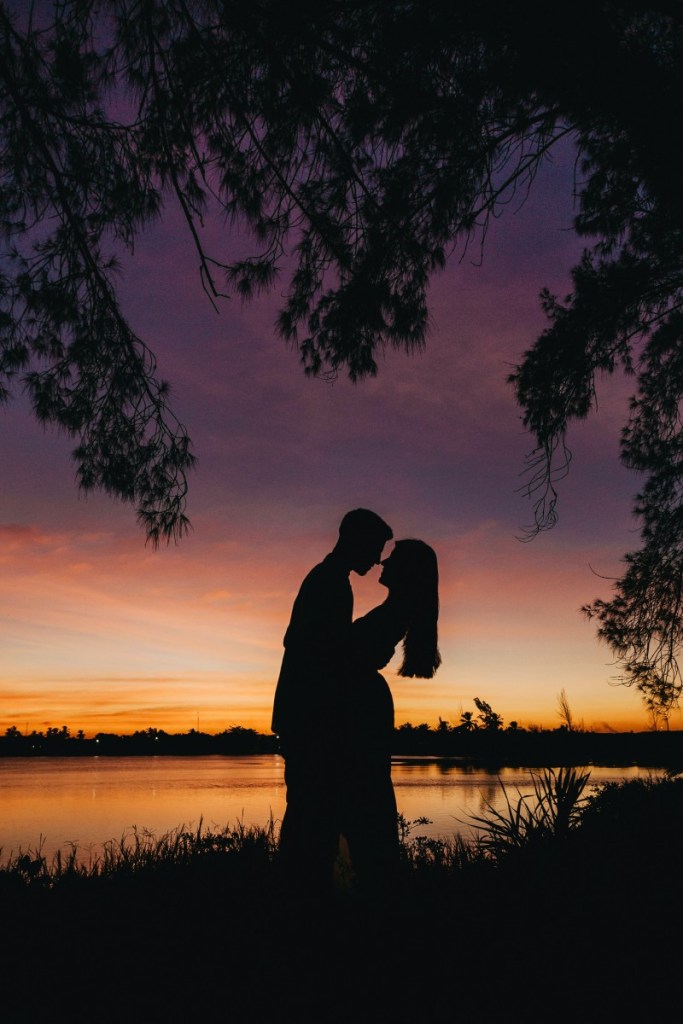 Casal abraçados quase se beijando em frente a um lago, está se pondo o sol, então só é possível ver a silhueta de sombra deles