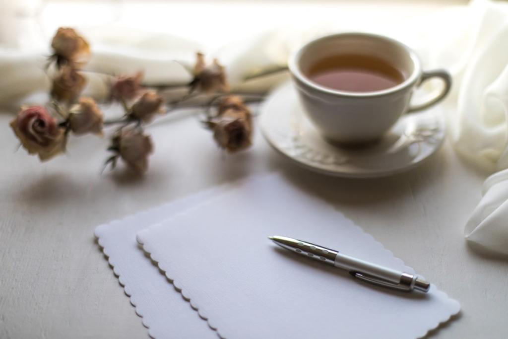 Em cima da mesa folhas, caneta, flores secas e um chá