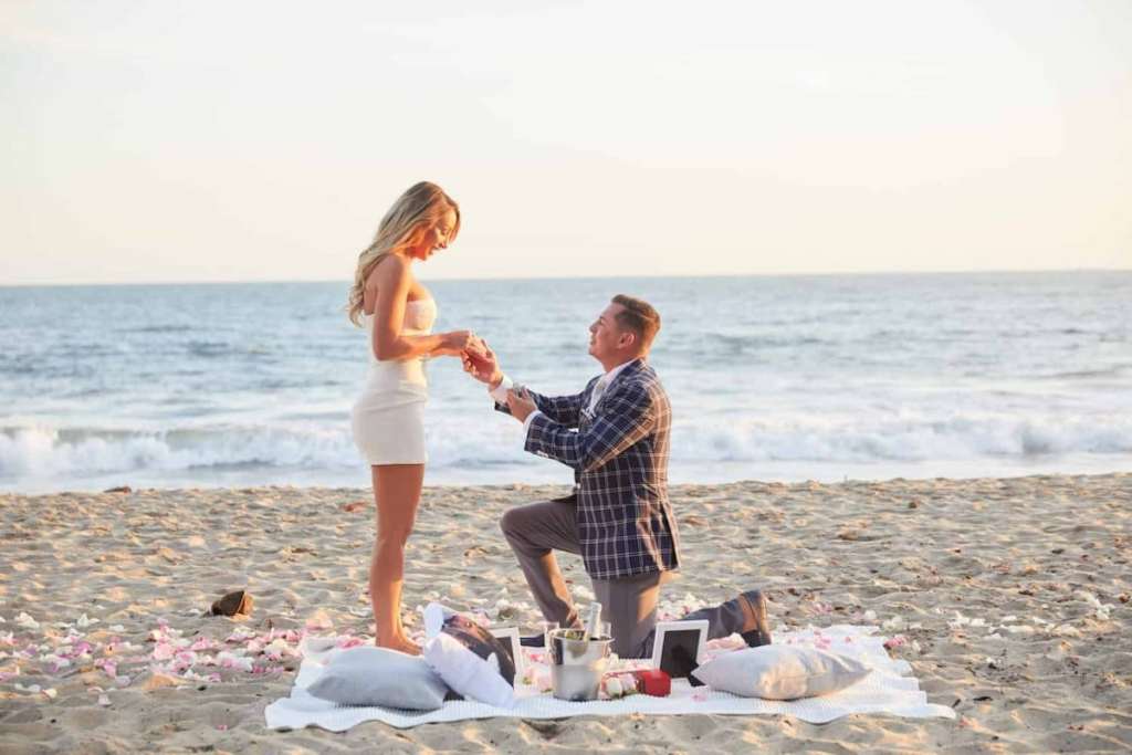 Homem pedindo mulher em casamento ajoelhado na praia.