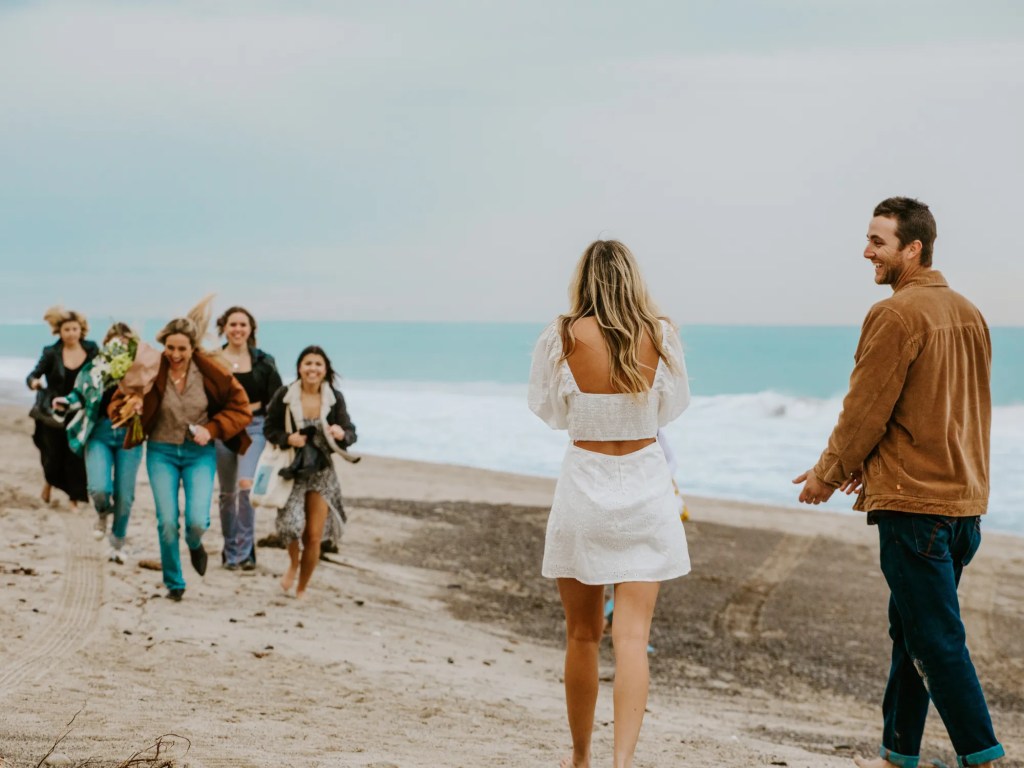 Homem pedindo mulher em casamento na praia, suas amigas chegam correndo