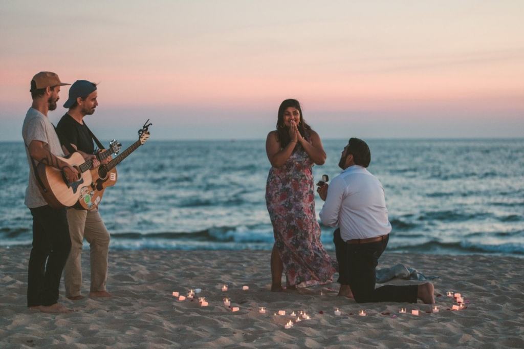 Homem pedindo mulher em casamento na praia, enquanto dupla de rapazes cantam com violão - como fazer o pedido de casamento na praia?