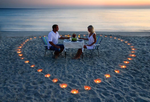 Casal jantando na praia, tem um coração feito de velas ao redor da mesa deles