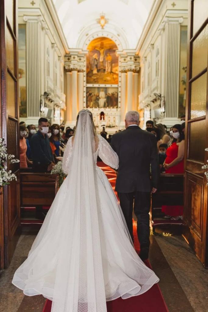 Noiva entrando no casamento segurando o braço do seu pai, os convidados estão em pé usando máscara
