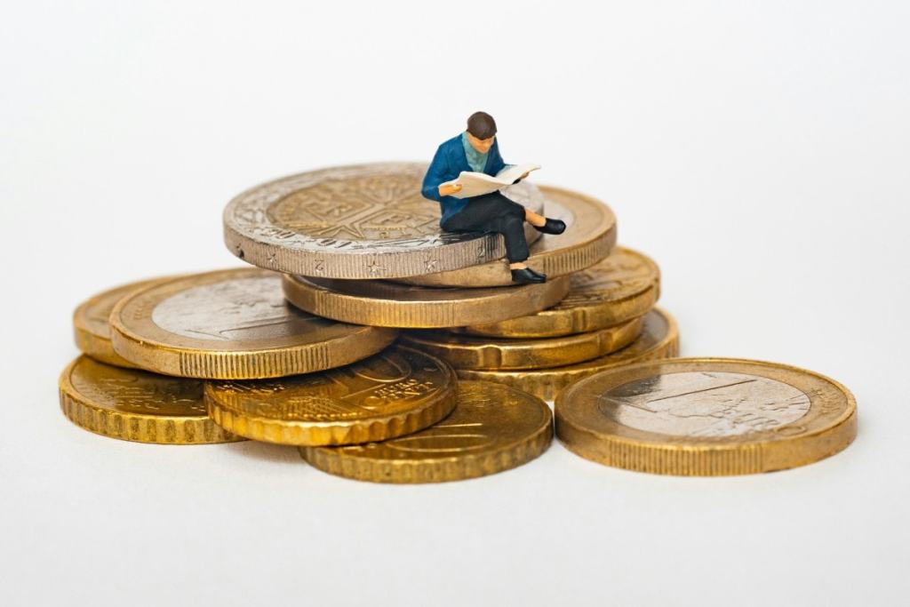 Algumas moedas e em bonequinho de um homem lendo jornal sentado em cima da moeda