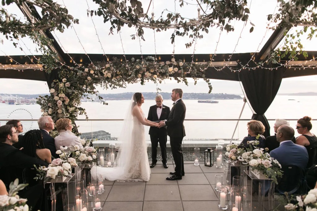 Noivos se casando em um rooftop de dia, eles estão de mãos dadas olhando um para o outro, e os convidados sentados - como fazer um casamento ao ar livre?