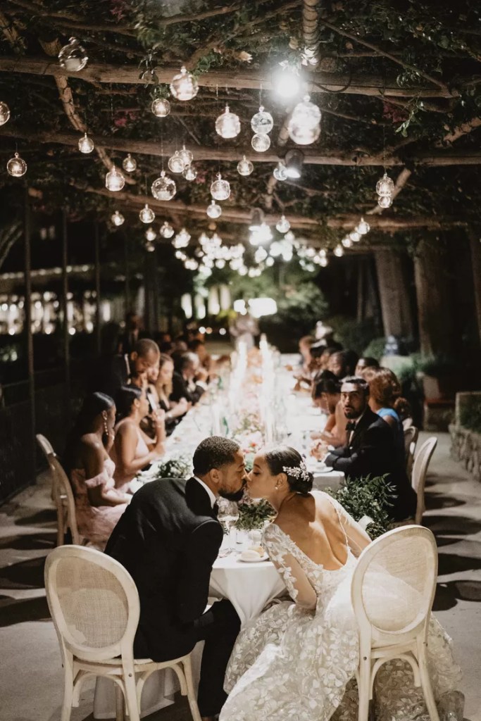 Casamento ao ar livre a noite, tem pequenos candelabros pendurados nos galhos que estão em cima da mesa principal, os noivos estão se beijando