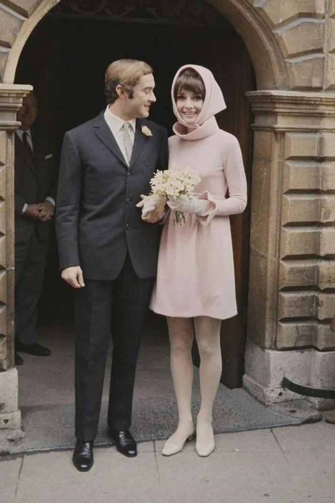 Vestido de noiva da audrey hepburn é curto e rosa, ela está parada ao lado do marido que olha para ela