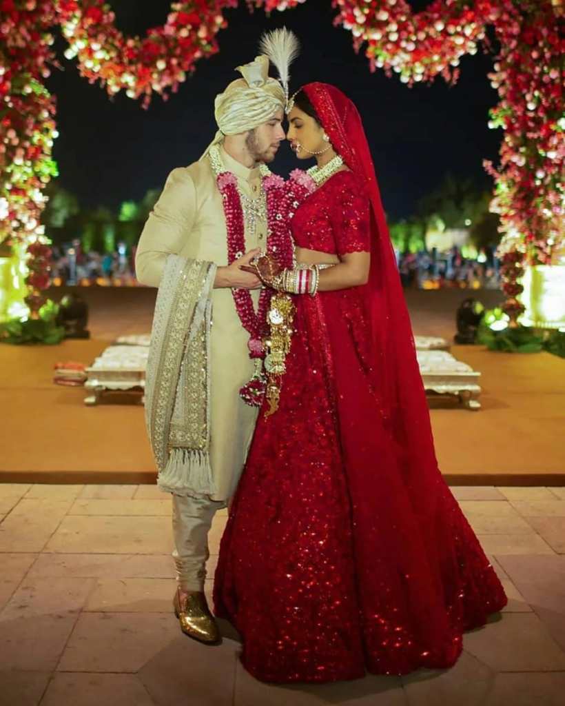Priyanka chopra e nick jonas em seu casamento, eles estão de mãos dadas e olhando um para o outro com rosto próximo, eles estão com roupas de casamento tradicionais hindu
