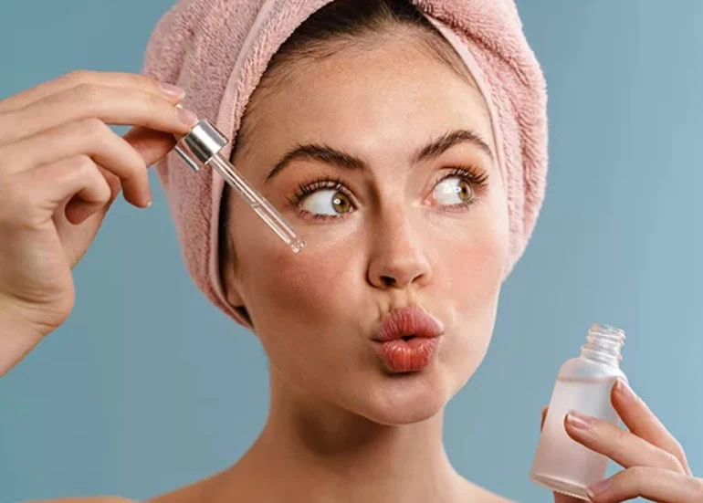 Mulher fazendo biquinho enquanto aplica sérum em gotas no rosto, ela está segurando o frasco na outra mão e possui uma toalha rosa no cabelo - rotinas de beleza pré-casamento