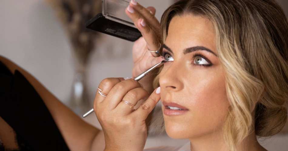 Maquiadora passando sombra em noiva. Imagem tem vista lateral do rosto da noiva.