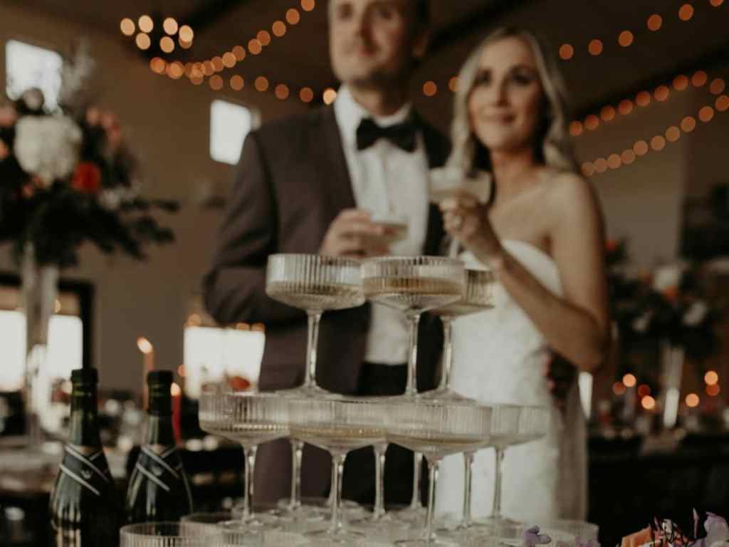 Noivos estão sorrindo desfocados no fundo enquanto seguram taças, na parte da frente da imagem tem uma torre de taças com champagne dentro