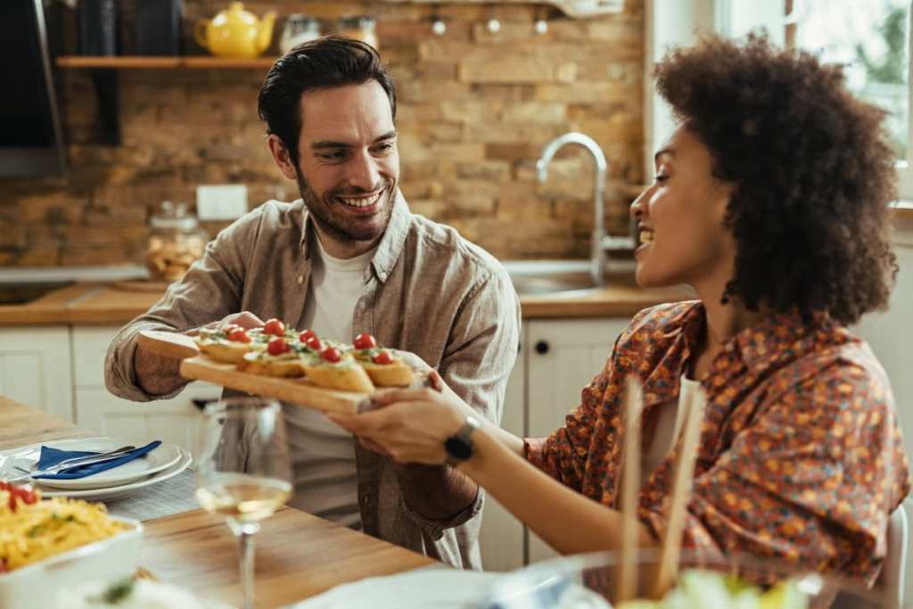 Homem e mulher em uma cozinha almoçando, ele está passando uma tábua com torradas para ela sorrindo