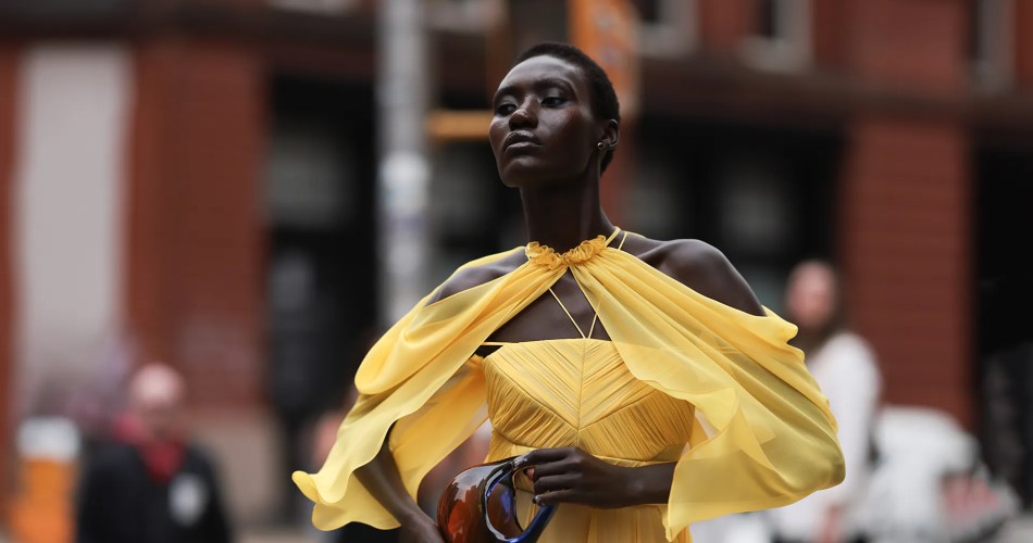 Mulher negra, com cabelo raspado, usando vestido longo amarelo com estola fina. Ela está andando na rua.