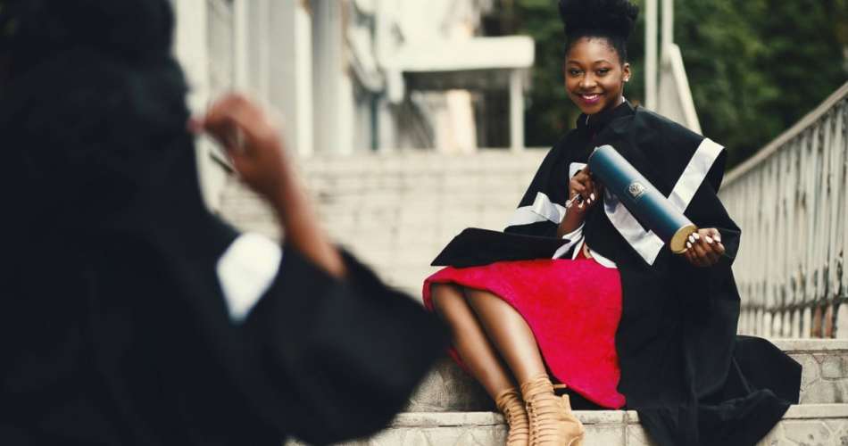 Mulher com vestido vermelho e beca preta de formatura e capelo, sentada em escada segurando diploma e posando para foto
