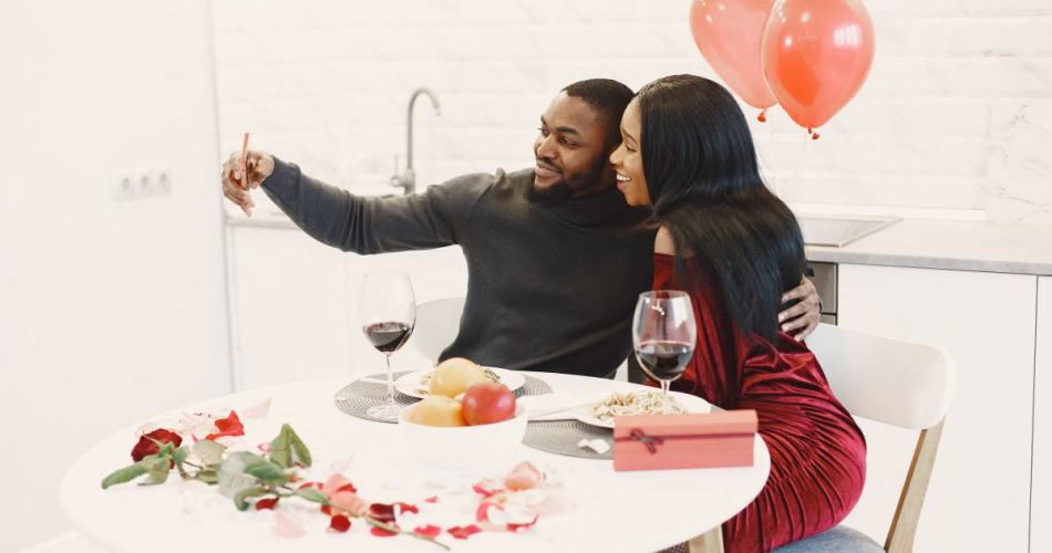 Casal sentado em mesa de jantar com pétalas de rosas e balões vermelhos, eles estão felizes e tirando uma selfie juntos.